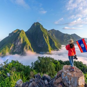 Asian girl holding Laos flag on top of Viewpoint of Nong Khiaw - a secret village in Laos. Stunning scenery of limestone cliff valley covered with fog
