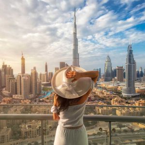 Woman with a white hat is standing on a balcony in front of the skyline from Dubai